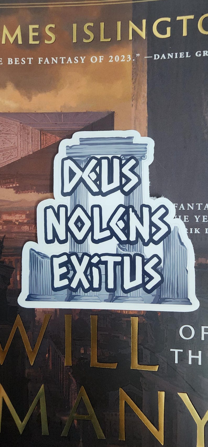 Puede incluir: Una pegatina blanca con la frase latina "Deus Nolens Exitus" en letras azules estilizadas. El dise&ntilde;o incorpora elementos arquitect&oacute;nicos como columnas y un templo, sobre un fondo de portada de libro.