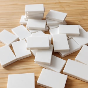 May include: A collection of small, white rectangular tiles scattered on a light brown wooden surface. The tiles are uniform in size and shape, with a smooth, matte finish. Some tiles are stacked, while others are spread out.