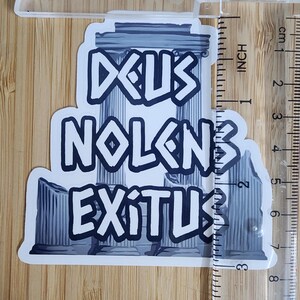 Puede incluir: Una pegatina con las palabras "DEUS NOLENS EXITUS" en una fuente estilizada, con un dise&ntilde;o de columna romana. La pegatina mide aproximadamente 7.6 cm de altura y est&aacute; colocada sobre una superficie de madera.