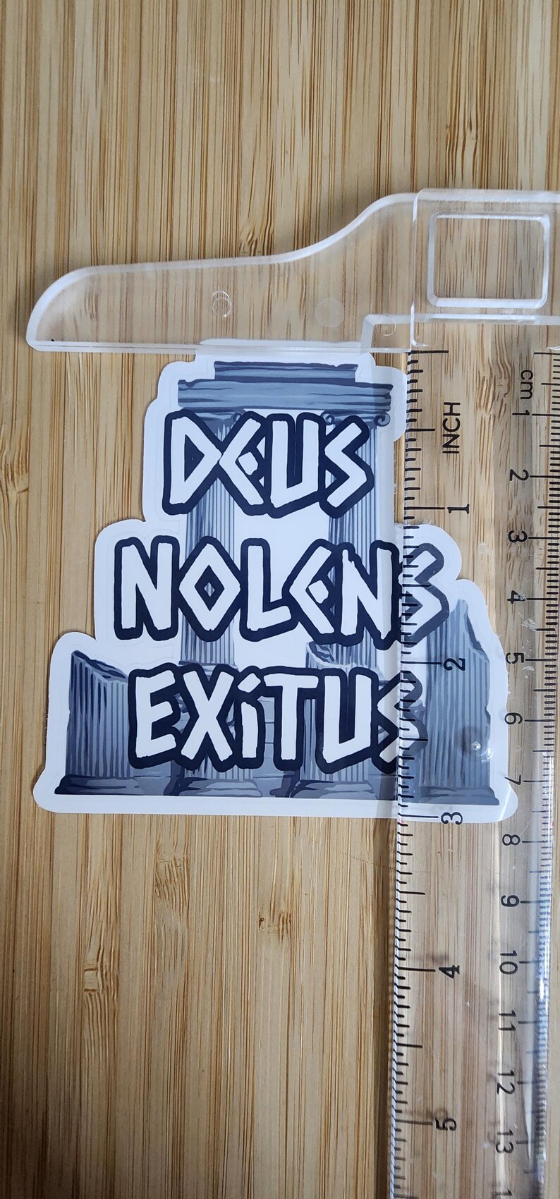 Puede incluir: Una pegatina con las palabras "DEUS NOLENS EXITUS" en una fuente estilizada, con un dise&ntilde;o de columna romana. La pegatina mide aproximadamente 7.6 cm de altura y est&aacute; colocada sobre una superficie de madera.