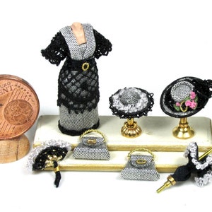 Peut inclure: Une vitrine miniature de maison de poupée présentant une robe noire et argentée, deux chapeaux noirs à bordure blanche, deux sacs à main argentés, un éventail noir et blanc et un parasol noir à bordure blanche. La vitrine comprend également une petite table blanche avec un vase de fleurs blanches.