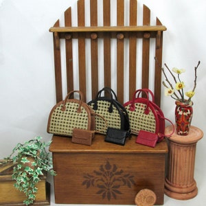 K&ouml;nnte beinhalten: Vier Miniatur-Handtaschen in verschiedenen Farben und Stilen, darunter Schwarz, Braun und Rot, werden auf einem Holzregal mit einem dekorativen Design pr&auml;sentiert. Das Regal ist an einer Holzstruktur befestigt, die einer Zaunstruktur &auml;hnelt. Eine kleine Topfpflanze mit gr&uuml;nen Bl&auml;ttern befindet sich im Vordergrund.