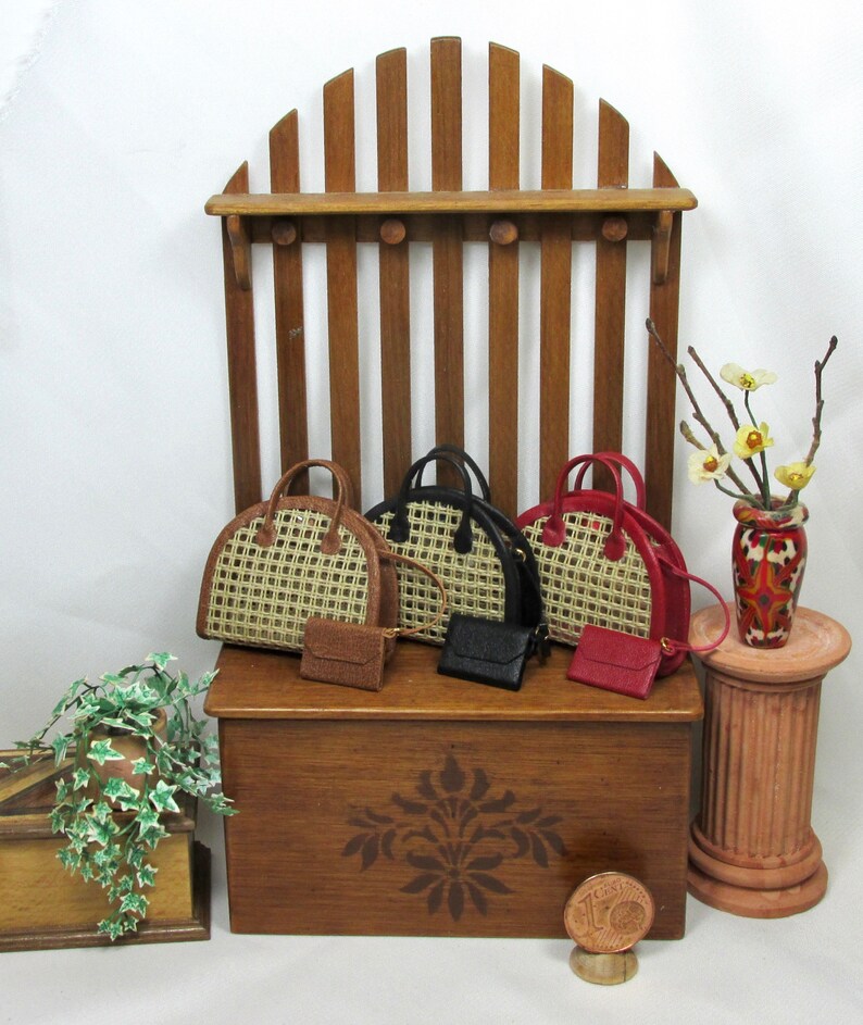 K&ouml;nnte beinhalten: Vier Miniatur-Handtaschen in verschiedenen Farben und Stilen, darunter Schwarz, Braun und Rot, werden auf einem Holzregal mit einem dekorativen Design pr&auml;sentiert. Das Regal ist an einer Holzstruktur befestigt, die einer Zaunstruktur &auml;hnelt. Eine kleine Topfpflanze mit gr&uuml;nen Bl&auml;ttern befindet sich im Vordergrund.
