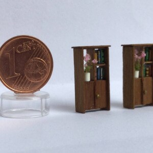 May include: Two miniature wooden bookcases with shelves and doors. Each bookcase has a small vase with pink flowers on the top shelf. The bookcases are painted brown and have a rustic look.