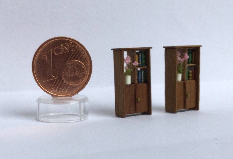 May include: Two miniature wooden bookcases with shelves and doors. Each bookcase has a small vase with pink flowers on the top shelf. The bookcases are painted brown and have a rustic look.
