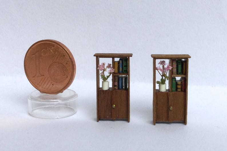 May include: Three miniature wooden bookcases with glass doors and shelves. Each bookcase has a small vase with pink flowers on the top shelf and books on the lower shelves.