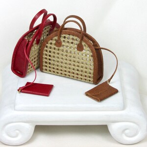 K&ouml;nnte beinhalten: Drei Miniatur-Handtaschen auf einem wei&szlig;en Sockel. Eine Handtasche ist aus rotem Leder mit einem geflochtenen Strohmuster. Die beiden anderen Handtaschen sind aus geflochtenem Stroh mit braunen Lederbes&auml;tzen.