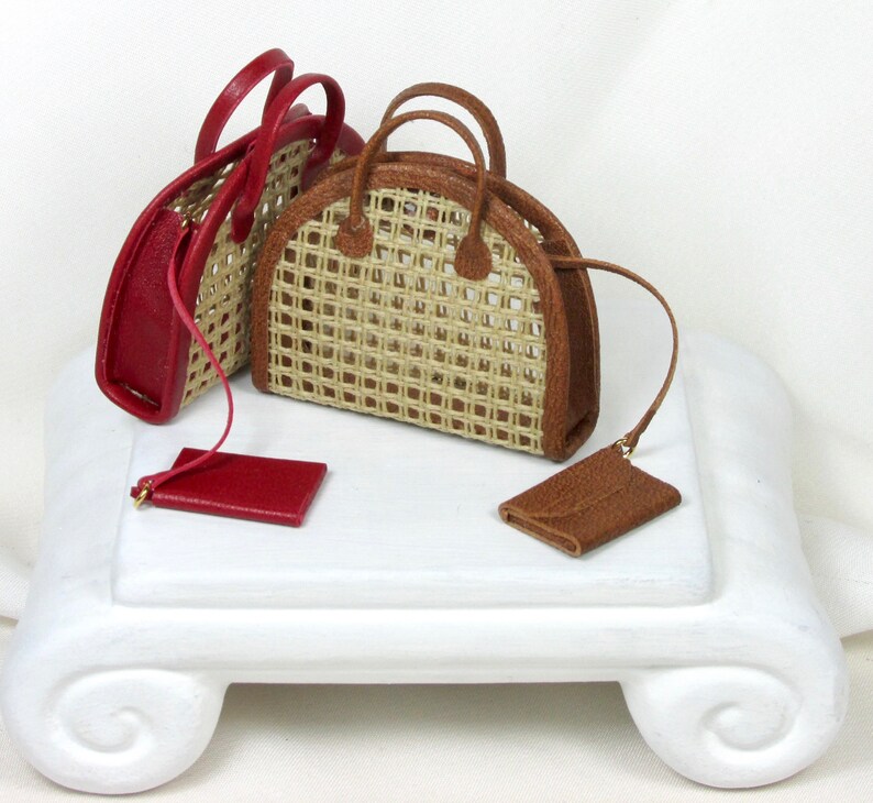 K&ouml;nnte beinhalten: Drei Miniatur-Handtaschen auf einem wei&szlig;en Sockel. Eine Handtasche ist aus rotem Leder mit einem geflochtenen Strohmuster. Die beiden anderen Handtaschen sind aus geflochtenem Stroh mit braunen Lederbes&auml;tzen.