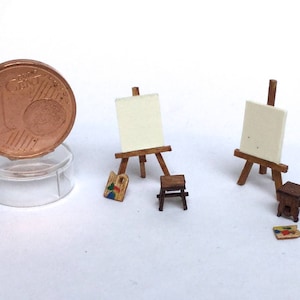 May include: Miniature artist's set featuring two wooden easels with blank canvases, small wooden stools, and paint palettes. A one Euro cent coin is included for scale, displayed on a clear plastic stand. The set is designed for dollhouses or craft projects.
