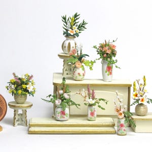 May include: A collection of miniature floral arrangements in white and floral patterned vases on a set of white shelves. The flowers are pink, yellow, and white.