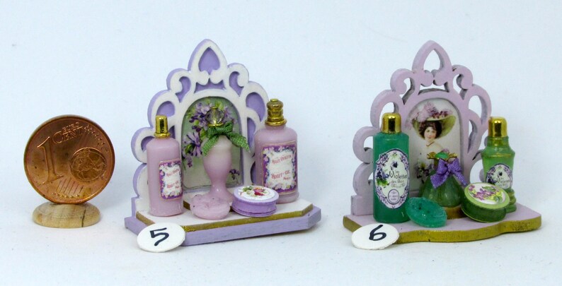May include: Two miniature bathroom vanity sets with decorative mirrors, shelves, and bottles of perfume. The set on the left has a purple and white color scheme with a floral design. The set on the right has a green and white color scheme with a floral design and a woman's portrait.