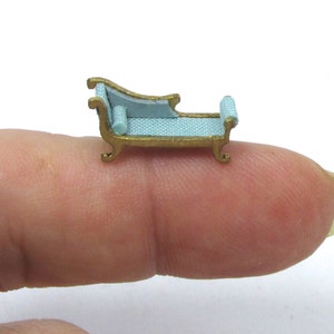May include: A miniature chaise lounge with a light blue fabric covering and gold-colored wooden frame. The chaise lounge is sitting on a finger.