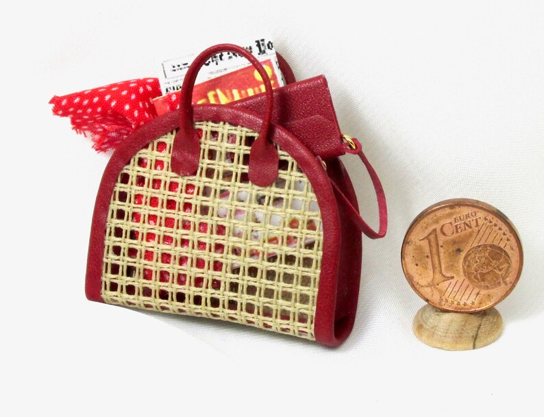K&ouml;nnte beinhalten: Eine rote geflochtene Handtasche mit einem braunen Ledergriff und einem rot-wei&szlig; gepunkteten Schal im Inneren. Die Tasche ist etwa so gro&szlig; wie eine Ein-Euro-M&uuml;nze.