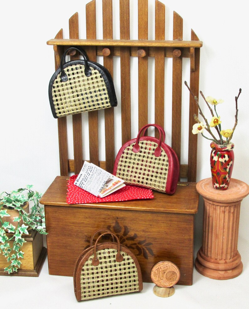 K&ouml;nnte beinhalten: Drei Miniatur-Korbtaschen mit Lederbesatz. Die Handtaschen werden auf einem Holzregal mit einem rot-wei&szlig; gepunkteten Stoff und einer Zeitung darauf ausgestellt. Die Handtaschen sind schwarz, rot und braun.