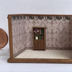 May include: A miniature dollhouse room with a wooden bookcase with glass doors and shelves filled with books. The bookcase is in front of a wallpapered wall with a floral pattern. The room is empty except for the bookcase.
