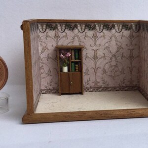 May include: A miniature dollhouse room with a wooden floor and a brown wooden bookcase with a floral arrangement and books. The walls are covered in a pink and white patterned wallpaper with a floral trim.