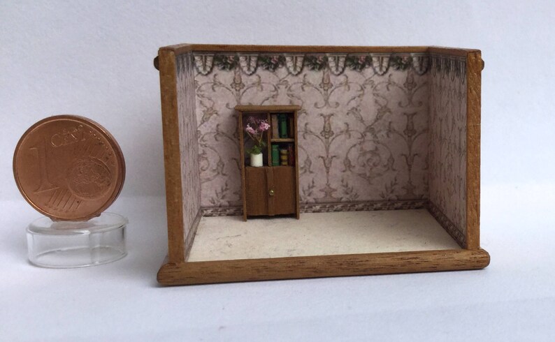 May include: A miniature dollhouse room with a wooden floor and a brown wooden bookcase with a floral arrangement and books. The walls are covered in a pink and white patterned wallpaper with a floral trim.