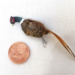 May include: A miniature pheasant figurine with detailed feathering in shades of brown, tan, and orange. The bird has a red and black head with a white collar. A one-cent coin is next to the bird for size reference.