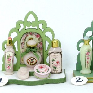 May include: Two miniature green wooden display stands with ornate designs, each holding a collection of miniature bottles of perfume and other toiletries. The bottles are decorated with floral patterns and have labels with text in French. The stands are on a white surface.
