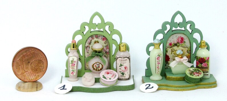 May include: Two miniature green wooden display stands with ornate designs, each holding a collection of miniature bottles of perfume and other toiletries. The bottles are decorated with floral patterns and have labels with text in French. The stands are on a white surface.