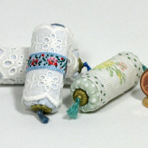 Może przedstawiać: Miniaturowe rolki materiału, niektóre owinięte białą koronką, inne we wzory kwiatowe lub w kropki. Każda rolka ma ozdobną mosiężną końcówkę i chwost. Dołączona jest moneta 1 eurocent jako skala.