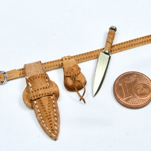 May include: A brown leather belt with a silver buckle, two knife sheaths, and a small pouch. The belt is decorated with stitching. A silver knife is attached to the belt.