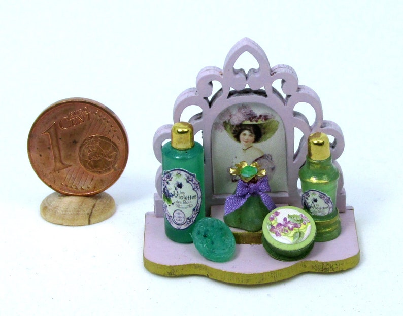May include: A miniature vanity set with a pink frame mirror, two green glass bottles with labels that say "Violettes", a green glass bottle with a purple ribbon, and a green soap dish with a floral design.