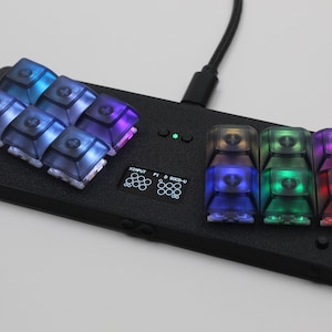 May include: A black rectangular gaming controller featuring two clusters of six translucent mechanical keycaps with integrated RGB lighting. A small central OLED screen displays the text XINPUT P1 D SOCD-U, accompanied by a single green LED indicator.