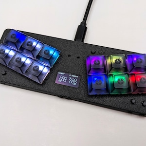 Könnte beinhalten: Eine schwarze Tastatur mit einem kleinen Bildschirm, auf dem der Text "INPUT 0 SOCD-IN" angezeigt wird. Die Tastatur hat 13 Tasten, von denen 7 transparent sind und mit blauem und violettem Licht beleuchtet werden, und 6 sind transparent und mit verschiedenen Farben beleuchtet, darunter Rot, Grün, Blau und Violett.