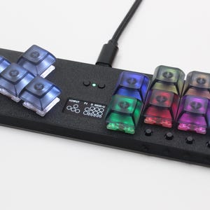 Op de afbeelding: Een zwarte rechthoekige controller met een groep van vier doorschijnende donkerblauwe toetsen aan de linkerkant en acht kleurrijke doorschijnende toetsen aan de rechterkant. Een klein scherm toont de tekst XINPUT P1 D 80CD-U met een groen indicatielampje.
