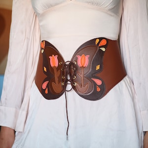 May include: A brown leather corset with a butterfly design. The corset has a lace-up front and is decorated with pink and orange flowers.