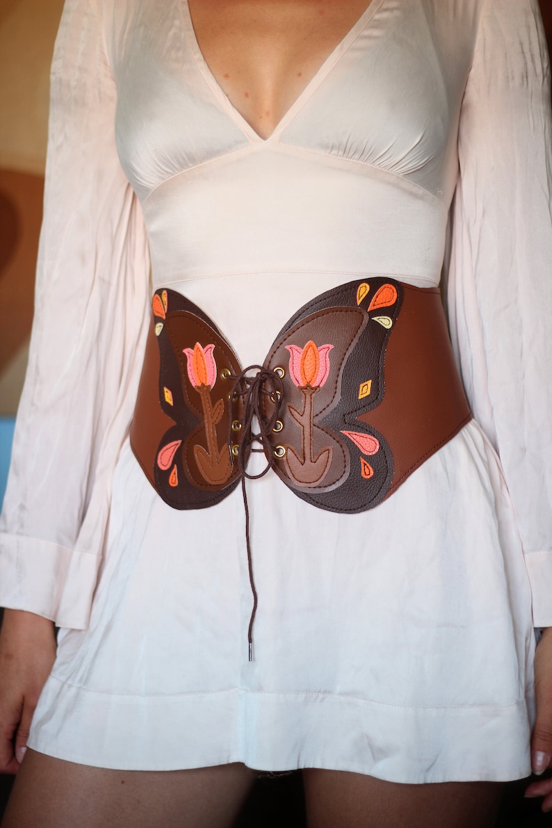 May include: A brown leather corset with a butterfly design. The corset has a lace-up front and is decorated with pink and orange flowers.