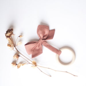 Peut inclure: Un hochet pour b&eacute;b&eacute; avec un anneau en bois et un n&oelig;ud rose poudr&eacute;. Le hochet est fait de tissu doux et de bois. Des fleurs s&eacute;ch&eacute;es sont dispos&eacute;es sur un fond blanc.
