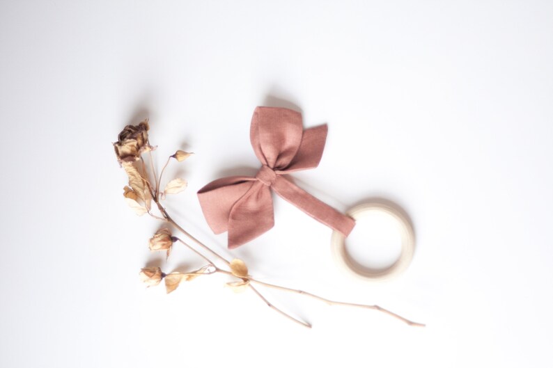 Peut inclure: Un hochet pour b&eacute;b&eacute; avec un anneau en bois et un n&oelig;ud rose poudr&eacute;. Le hochet est fait de tissu doux et de bois. Des fleurs s&eacute;ch&eacute;es sont dispos&eacute;es sur un fond blanc.