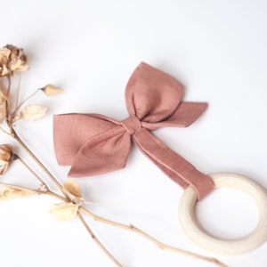 Peut inclure: Un hochet pour b&eacute;b&eacute; de couleur rose poudr&eacute;, avec un anneau en bois et un n&oelig;ud en tissu. Le n&oelig;ud est fix&eacute; &agrave; l'anneau par une bande de tissu assortie. Des fleurs s&eacute;ch&eacute;es sont en arri&egrave;re-plan.
