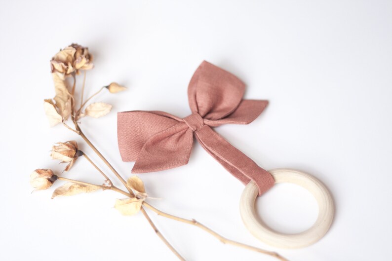 Peut inclure: Un hochet pour b&eacute;b&eacute; de couleur rose poudr&eacute;, avec un anneau en bois et un n&oelig;ud en tissu. Le n&oelig;ud est fix&eacute; &agrave; l'anneau par une bande de tissu assortie. Des fleurs s&eacute;ch&eacute;es sont en arri&egrave;re-plan.