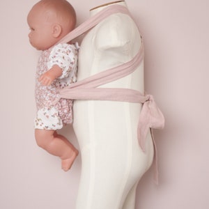 Peut inclure: Un porte-b&eacute;b&eacute; rose avec une poup&eacute;e v&ecirc;tue d'une robe &agrave; fleurs. Le porte-b&eacute;b&eacute; est attach&eacute; autour d'un mannequin blanc.