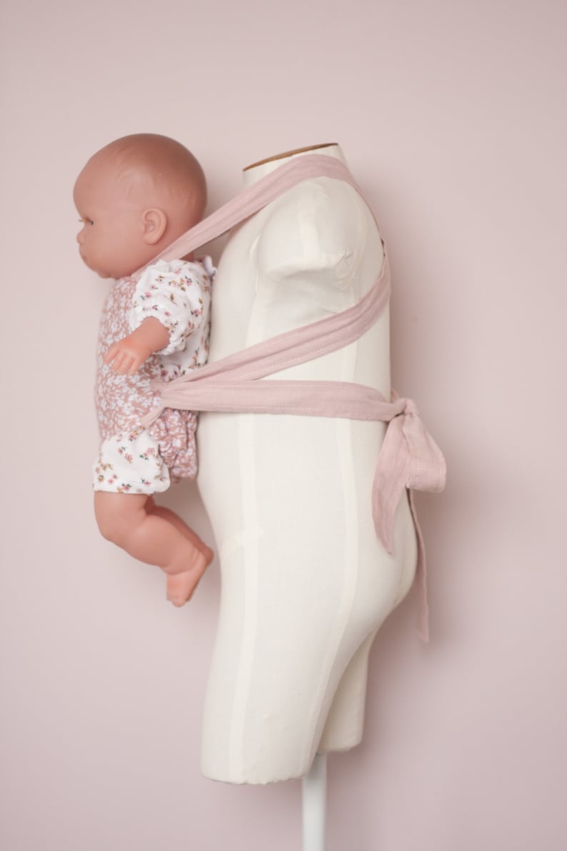 Peut inclure: Un porte-b&eacute;b&eacute; rose avec une poup&eacute;e v&ecirc;tue d'une robe &agrave; fleurs. Le porte-b&eacute;b&eacute; est attach&eacute; autour d'un mannequin blanc.