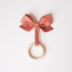 Peut inclure: Un jouet pour b&eacute;b&eacute; compos&eacute; d'un anneau en bois et d'un n&oelig;ud papillon en tissu de couleur corail. Le n&oelig;ud est fix&eacute; &agrave; l'anneau par une bande de tissu assortie. Le jouet est pos&eacute; sur un fond blanc.