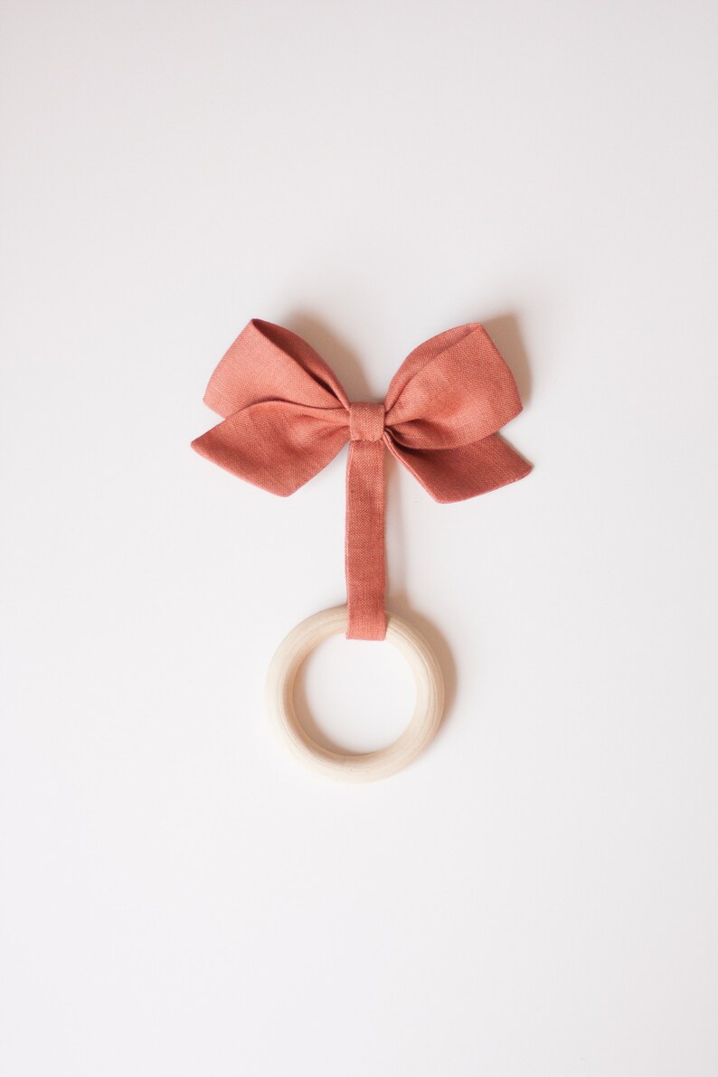 Peut inclure: Un jouet pour b&eacute;b&eacute; compos&eacute; d'un anneau en bois et d'un n&oelig;ud papillon en tissu de couleur corail. Le n&oelig;ud est fix&eacute; &agrave; l'anneau par une bande de tissu assortie. Le jouet est pos&eacute; sur un fond blanc.