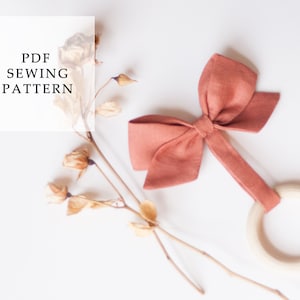 Peut inclure: Un hochet pour b&eacute;b&eacute; fait &agrave; la main, avec un n&oelig;ud corail et un anneau en bois. Le hochet est pr&eacute;sent&eacute; avec des fleurs s&eacute;ch&eacute;es et le texte "PDF SEWING PATTERN" sur fond blanc. Id&eacute;al comme cadeau de naissance.