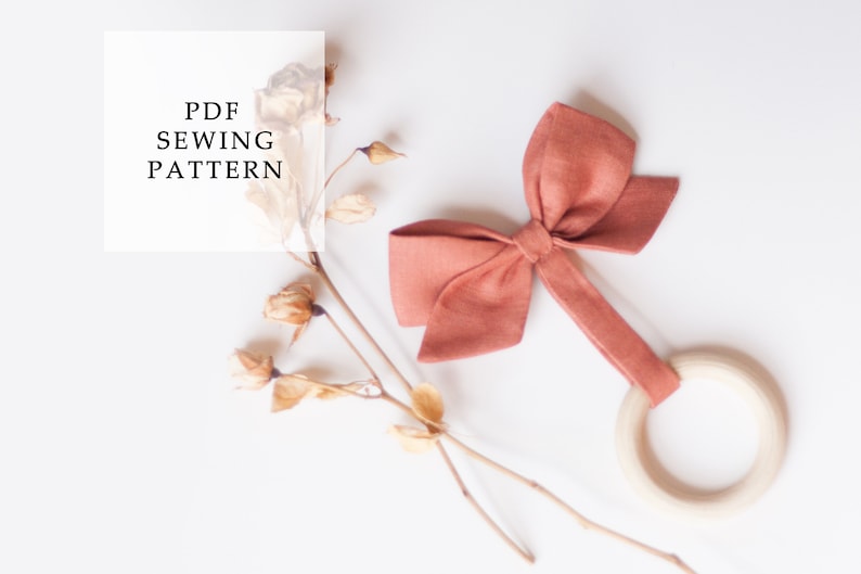 Peut inclure: Un hochet pour b&eacute;b&eacute; fait &agrave; la main, avec un n&oelig;ud corail et un anneau en bois. Le hochet est pr&eacute;sent&eacute; avec des fleurs s&eacute;ch&eacute;es et le texte "PDF SEWING PATTERN" sur fond blanc. Id&eacute;al comme cadeau de naissance.