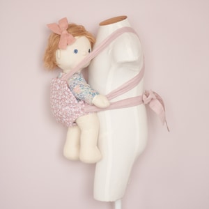 Peut inclure: Une poup&eacute;e b&eacute;b&eacute; rose avec une robe &agrave; fleurs est port&eacute;e dans un porte-b&eacute;b&eacute; rose sur un mannequin blanc.
