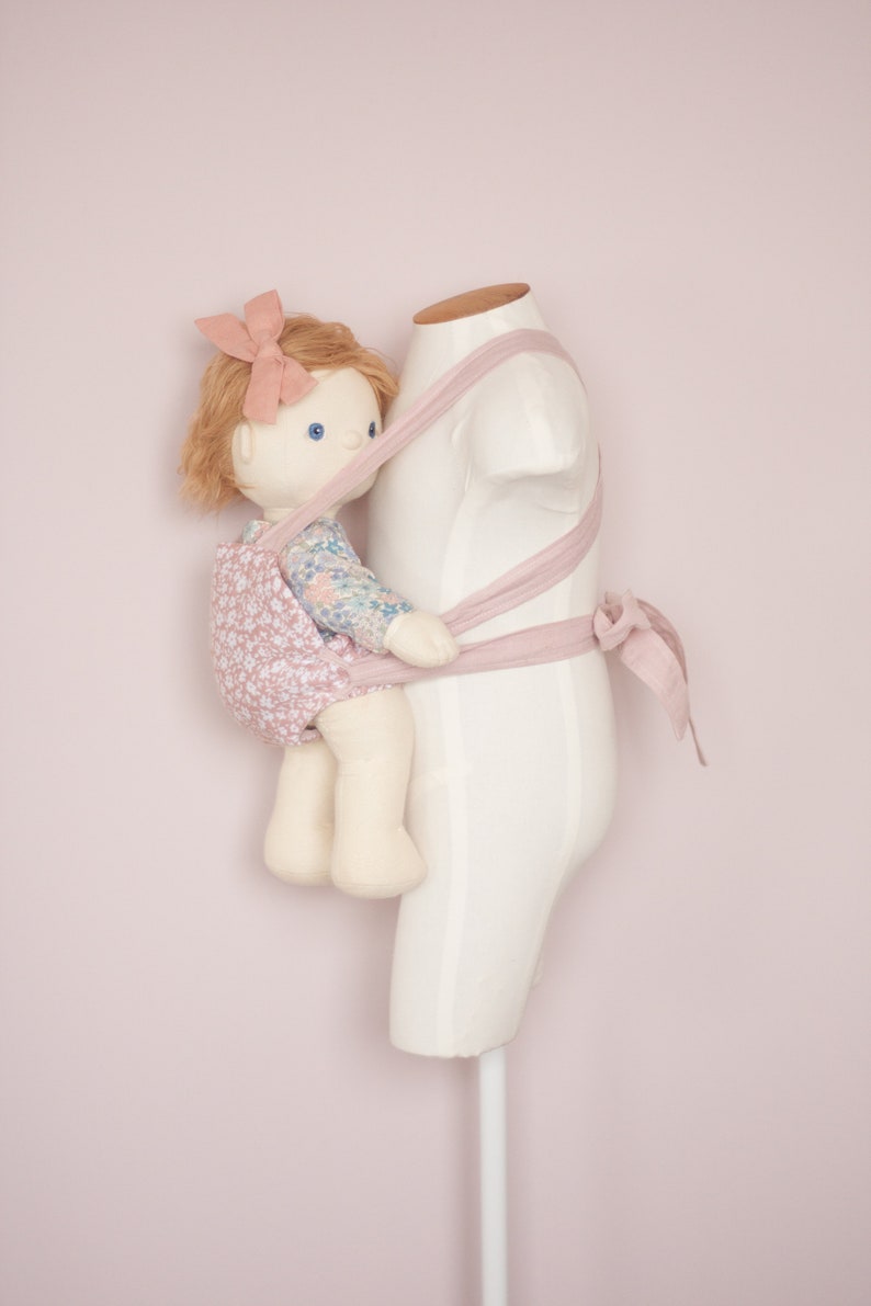 Peut inclure: Une poup&eacute;e b&eacute;b&eacute; rose avec une robe &agrave; fleurs est port&eacute;e dans un porte-b&eacute;b&eacute; rose sur un mannequin blanc.