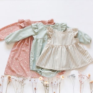 Op de afbeelding: Drie kinderkledingstukken: een oudroze jurk met lange mouwen en een wit bloemenpatroon, een saliegroene romper met lange mouwen en ruches op de schouders, en een mouwloze beige en wit gestreepte jurk met ruches op de schouders.