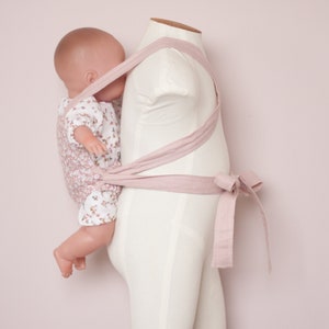 Peut inclure: Un porte-b&eacute;b&eacute; rose en tissu doux est pr&eacute;sent&eacute; sur un mannequin blanc. Le porte-b&eacute;b&eacute; est nou&eacute; &agrave; la taille et poss&egrave;de des bretelles qui passent par-dessus les &eacute;paules. Un poupon est port&eacute; dans le porte-b&eacute;b&eacute;.