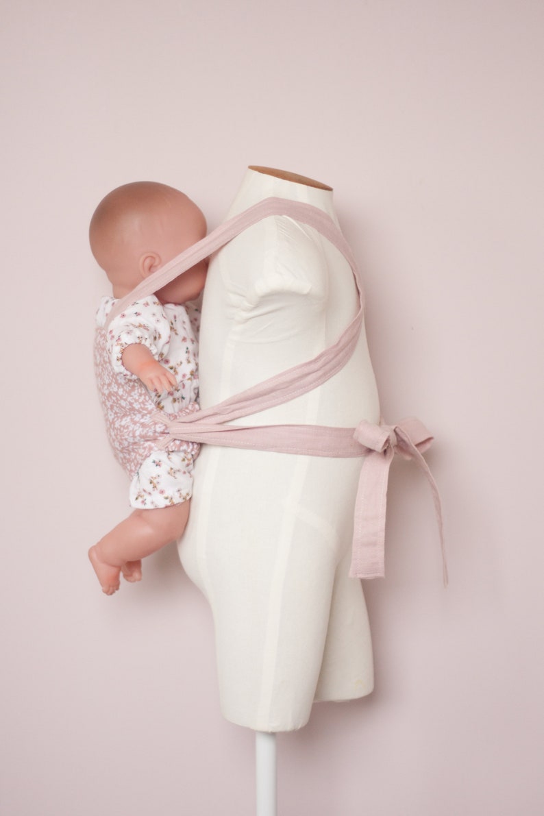 Peut inclure: Un porte-b&eacute;b&eacute; rose en tissu doux est pr&eacute;sent&eacute; sur un mannequin blanc. Le porte-b&eacute;b&eacute; est nou&eacute; &agrave; la taille et poss&egrave;de des bretelles qui passent par-dessus les &eacute;paules. Un poupon est port&eacute; dans le porte-b&eacute;b&eacute;.