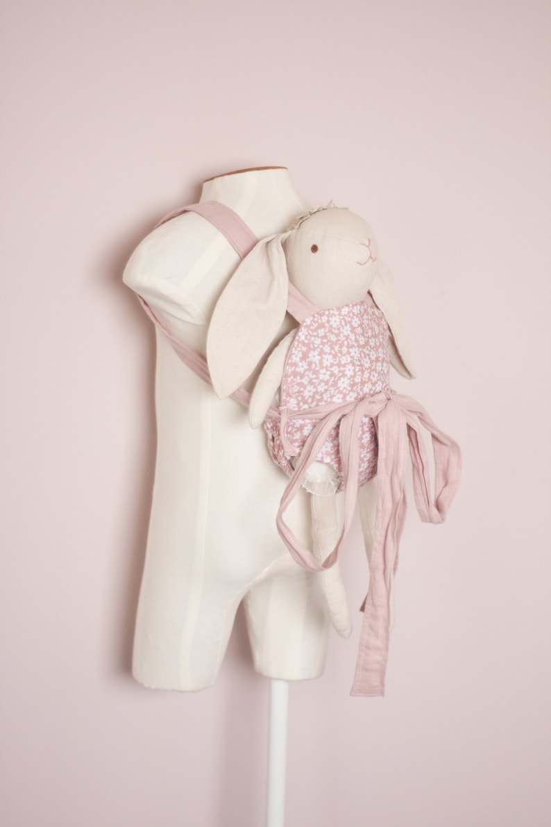 Peut inclure: Un porte-b&eacute;b&eacute; floral rose et blanc avec une poup&eacute;e lapin attach&eacute;e. Le porte-b&eacute;b&eacute; a de longues attaches qui peuvent &ecirc;tre nou&eacute;es autour de la taille.