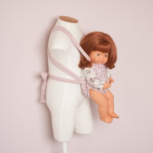 Peut inclure: Un porte-b&eacute;b&eacute; rose avec une poup&eacute;e v&ecirc;tue d'une robe &agrave; fleurs blanches et roses. La poup&eacute;e a les cheveux bruns et est assise dans le porte-b&eacute;b&eacute;.