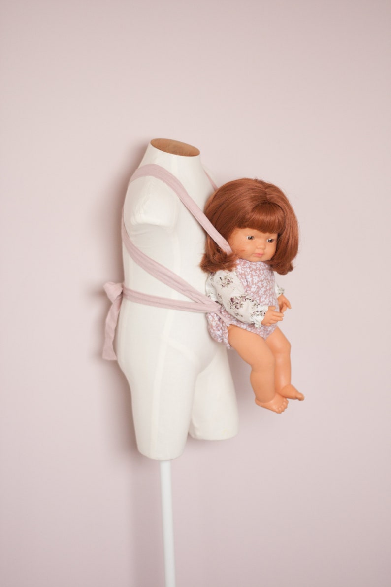 Peut inclure: Un porte-b&eacute;b&eacute; rose avec une poup&eacute;e v&ecirc;tue d'une robe &agrave; fleurs blanches et roses. La poup&eacute;e a les cheveux bruns et est assise dans le porte-b&eacute;b&eacute;.